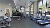 This single-level fitness center is thoughtfully designed to support a well-rounded workout experience. The space features a dark rubber flooring that provides both durability and cushioning underfoot, making it suitable for a variety of exercise activities. Along one wall, floor-to-ceiling mirrors visually expand the room while allowing users to monitor their form during workouts. A comprehensive free weight section lines the mirrored wall, offering a full rack of dumbbells alongside a dedicated weight bench. The cardio area is positioned near large arched windows that frame views of lush outdoor greenery, flooding the space with natural light and creating an open, airy atmosphere. Equipment includes elliptical machines and treadmills, catering to a range of cardiovascular training preferences. A wall-mounted flat-screen television is positioned for easy viewing from multiple workout stations, and a ceiling fan overhead helps maintain comfortable airflow throughout the room. The vaulted ceiling adds a sense of openness, contributing to the overall welcoming and functional ambiance of the fitness facility.