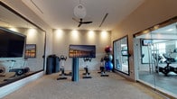 This single-floor fitness studio is thoughtfully designed to support a variety of cardio and group exercise activities. The room centers around a row of spin bikes arranged to face a large wall-mounted flat-screen display, making it well-suited for guided cycling classes or individual workout sessions. A decorative photographic print of a pier at dusk adds a coastal aesthetic touch to the main wall, contributing to a calm yet energizing ambiance.

The space is well-lit through a combination of recessed ceiling spotlights and a distinctive globe pendant light, ensuring even illumination throughout. A ceiling fan overhead aids in air circulation, keeping the environment comfortable during high-intensity workouts. The neutral-toned carpet flooring provides a practical, cushioned surface underfoot.

A collection of stability and medicine balls in varying sizes is neatly stacked in one corner, offering additional versatility for strength and balance training. A large framed window allows natural light to filter in while providing a view to an adjacent area, enhancing the open feel of the room. A wall-mounted mirror further amplifies the sense of space and supports proper form during exercise.