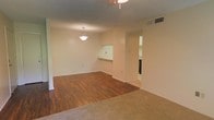 This single-story space presents an open, functional layout with a seamless transition between the carpeted and hardwood-style flooring zones, visually distinguishing the living and dining areas. The living area features warm-toned carpet, while the adjoining dining space is finished with rich, dark laminate or hardwood flooring, adding contrast and visual interest to the neutral interior. Walls are painted in a soft, warm white that contributes to a bright and airy atmosphere throughout.

A pendant light fixture hangs above the dining area, providing focused illumination, while a ceiling fan is visible in the adjacent space, supporting airflow and comfort. A pass-through window connects the main living area to what appears to be the kitchen, enhancing the open feel and facilitating ease of communication between spaces. A ceiling vent indicates a central HVAC system is in place. A doorway leads to an additional room, partially visible in the background and appearing to include a bathroom or utility area. The overall design is clean and understated, with minimal architectural ornamentation, offering a versatile canvas well-suited to a variety of interior styles and furniture arrangements.