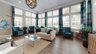 This well-appointed single-floor community space showcases a cohesive coastal design aesthetic defined by a rich palette of teal, navy, and soft gray tones set against warm hardwood-style flooring throughout. The main lounge area is a true centerpiece, featuring floor-to-ceiling windows that flood the room with natural light and frame outdoor views of a resort-style pool. Plush tufted teal wingback chairs, a neutral slipcovered sofa, and a rustic chandelier create an inviting and refined atmosphere. A secondary seating nook offers a more intimate setting, furnished with turquoise accent chairs and ocean-themed wall art, ideal for quiet conversation or relaxation. The space transitions smoothly into a well-equipped kitchenette featuring gray shaker-style cabinetry, granite countertops, under-cabinet lighting, and stainless steel appliances, providing functional amenities for residents or visitors. Near the main entry, a welcoming reception area is appointed with teal lounge chairs, patterned window valances, and French doors that open to the outdoors. Throughout the space, curated artwork, lush potted greenery, and thoughtful decorative accents reinforce the coastal theme, resulting in a polished, resort-inspired environment designed for both comfort and community engagement.