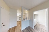 This single-floor unit offers a clean, functional layout with consistent wood-look flooring throughout, lending warmth and visual continuity across all areas. The kitchen is well-equipped with white shaker-style cabinetry extending to the ceiling for ample storage, white countertops, a double-basin stainless steel sink, and a gas range with an over-the-range microwave. A pass-through opening with a built-in shelf connects the kitchen to the adjacent living area, creating an open and communicative layout. The living area is bright and spacious, featuring a window unit for climate control and a large window that allows natural light to fill the room. A ceiling fan with integrated lighting is found in one of the rooms, adding both comfort and illumination. The bedroom areas are generously sized with neutral walls and textured ceilings, offering a calm and versatile canvas. A dedicated closet with built-in shelving provides practical storage. The space also features a bathroom accessible from the bedroom corridor, with white cabinetry visible from the hallway. Throughout the unit, clean white baseboards, smooth neutral-toned walls, and textured ceilings contribute to a cohesive and tidy aesthetic.