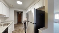 This 1-bedroom, 1-bathroom unit is thoughtfully laid out across a single floor, offering a comfortable and functional living environment. The galley-style kitchen is a standout feature, showcasing crisp white shaker cabinets, light granite-style countertops, wood-look flooring, and a full suite of appliances including a stainless steel refrigerator, electric range, and dishwasher. A round flush-mount ceiling light brightens the space efficiently. The living area is generously sized, finished with soft neutral carpet and clean white walls that create a bright, open atmosphere. A doorway connects the living space to the entry hallway, which is accented by a ceiling fan and a warm wood-tone front door. The bedroom continues the carpeted flooring and features a window with an integrated wall air conditioning unit and baseboard heating, ensuring year-round climate comfort along with views of the surrounding greenery. The bathroom is clean and straightforward, equipped with a full tub-shower combination, toilet, vanity with mirror, and baseboard heating. Throughout the unit, the neutral color palette and practical finishes provide a move-in ready living space suited for everyday comfort.