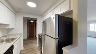 This 1-bedroom, 1-bathroom unit is thoughtfully laid out across a single floor, offering a comfortable and functional living environment. The galley-style kitchen is a standout feature, showcasing crisp white shaker cabinets, light granite-style countertops, wood-look flooring, and a full suite of appliances including a stainless steel refrigerator, electric range, and dishwasher. A round flush-mount ceiling light brightens the space efficiently. The living area is generously sized, finished with soft neutral carpet and clean white walls that create a bright, open atmosphere. A doorway connects the living space to the entry hallway, which is accented by a ceiling fan and a warm wood-tone front door. The bedroom continues the carpeted flooring and features a window with an integrated wall air conditioning unit and baseboard heating, ensuring year-round climate comfort along with views of the surrounding greenery. The bathroom is clean and straightforward, equipped with a full tub-shower combination, toilet, vanity with mirror, and baseboard heating. Throughout the unit, the neutral color palette and practical finishes provide a move-in ready living space suited for everyday comfort.