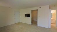 This single-floor space is designed with a straightforward, functional layout that maximizes usability. The main living area is finished with wall-to-wall neutral carpet, providing a soft underfoot feel throughout. White walls keep the space bright and adaptable to a variety of furnishing styles. A notable architectural detail is the pass-through window cut into the shared wall between the living area and the kitchen, allowing visibility and convenient access between the two spaces. Through this opening, kitchen appliances including a microwave are visible, suggesting a compact but practical kitchen setup. A doorway adjacent to the pass-through provides direct access to the kitchen and connects to a further adjoining room. A solid white entry door on the opposite wall offers a secondary access point, adding to the overall flow of the layout. Overhead, a ceiling-mounted HVAC vent indicates a central climate control system. The space is clean, unfurnished, and move-in ready, presenting a blank canvas well-suited for personalization. The overall ambiance is simple and utilitarian, emphasizing practicality and ease of use.
