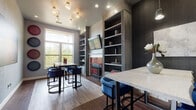 The Nook in Oxford, Mississippi is a thoughtfully designed single-floor gathering space that blends contemporary style with warm, inviting details. The main lounge area is anchored by a striking fireplace adorned with vibrant red subway tile and a wall-mounted television framed by floor-to-ceiling built-in shelving in a sophisticated gray finish. The shelving provides both decorative and functional display space, adding depth and character to the room. Seating throughout features plush blue velvet chairs paired with sleek marble-top tables supported by minimalist black metal frames, creating a refined yet comfortable atmosphere. The flooring transitions between rich dark tile and warm wood-toned planks, adding visual interest and defining distinct zones within the space. Large windows flood the lounge with natural light and offer views of the surrounding greenery, enhancing the open and airy feel. Decorative pendant lighting with globe fixtures adds a touch of elegance overhead. A wide corridor with textured dark accent walls connects the lounge to additional rooms, including a more intimate nook with its own built-in cabinetry, pendant lighting, and cozy seating arrangement. Throughout, the design palette of deep charcoals, rich blues, and warm wood tones creates a cohesive, upscale ambiance ideal for socializing or focused gatherings.