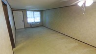 This single-floor space presents a straightforward, functional layout with a clean, open feel. The room features wall-to-wall carpeting in a neutral beige tone, providing a soft underfoot surface throughout. The walls are finished with a subtle textured wallcovering in muted green-beige hues, accented by dark baseboards and crown molding that add modest architectural definition. A textured ceiling completes the interior finish.

Natural light enters the space through a window fitted with vertical blinds, offering adjustable light control. Beneath the window sits a wall-mounted HVAC unit, providing both heating and cooling directly within the room. A doorway with a standard door is positioned along one wall, facilitating access to adjacent areas. The overall ambiance is practical and understated, with neutral tones throughout creating a versatile backdrop suitable for a variety of uses. The open floor plan allows for flexible furniture arrangement, making efficient use of the available space.