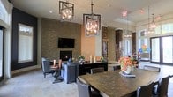 This thoughtfully designed single-floor space seamlessly blends comfort and sophistication through its open, flowing layout. The lounge area serves as a focal point, anchored by a floor-to-ceiling stacked stone accent wall that frames both a fireplace and a wall-mounted television, creating a warm and inviting gathering spot. Plush gray seating and refined accent furniture complete the relaxed yet polished atmosphere.

A dining area with a richly textured wood table and upholstered seating sits adjacent to the lounge, defined by a series of dramatic geometric pendant chandeliers that cast warm, ambient light throughout the space. The pendants serve as a striking architectural detail, tying the zones together visually.

Decorative mosaic tile columns add depth and texture, complementing the warm earth-toned walls and large-format tile flooring that runs throughout. A dark cabinetry console with display accessories provides both function and visual interest between the two areas.

Natural light filters in through doors at the rear of the space, hinting at an outdoor area beyond. Overall, the space exudes a contemporary, upscale ambiance with layered textures, warm lighting, and carefully curated design elements that make it both functional and visually engaging.
