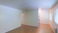 This single-floor space offers a clean, functional layout designed for comfortable everyday living. The main living area is bright and open, featuring warm hardwood-style flooring that extends throughout, creating a cohesive and inviting atmosphere. Crisp white walls reflect light effectively, contributing to a fresh and airy feel across the space. A centrally positioned HVAC vent ensures climate control throughout the living area. The space flows naturally into a partially open kitchen, which is visible through a pass-through opening. The kitchen features white cabinetry and a ceiling-mounted light fixture, providing practical illumination. The countertops offer a subtle contrasting tone, adding a modest design detail to the cooking area. The overall layout is straightforward and well-organized, with clear sightlines between the living and kitchen areas that enhance the sense of openness. Clean baseboards and neutral finishes throughout contribute to a well-maintained and move-in ready presentation. The single-level design ensures easy navigation and accessibility, making the space both practical and welcoming.