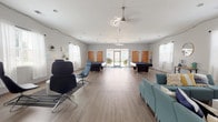 Located in Carrollton, GA, this single-floor community lounge is a well-appointed gathering space designed for both relaxation and recreation. The open layout is anchored by light gray wood-look flooring that runs throughout, complementing the soft gray-white walls and creating a clean, modern aesthetic.

The space is divided into several distinct functional zones. A lounge area is furnished with teal upholstered chairs and sofas arranged around a white coffee table, with a wall-mounted flat-screen TV and a shelving unit stocked with board games — ideal for casual gatherings. Adjacent to this, sleek black high-back swivel chairs are positioned on a patterned area rug, providing a more contemporary seating option.

Toward one end of the room, two billiard tables are prominently featured beneath ceiling fans and pendant lighting, with glass doors leading to an outdoor view and filling the space with natural light. Sheer white curtains frame multiple windows along the walls, keeping the interior bright and airy.

A dining or collaborative work area includes a long white high-top table surrounded by modern gray counter stools with yellow seat accents, set atop a textured area rug. Decorative string lights trace the upper walls, adding a warm, inviting ambiance throughout the entire space.