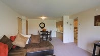 This single-floor residence is designed with an open-concept layout that seamlessly connects the living, dining, and kitchen areas into one cohesive and functional space. The living area features plush carpeting in a neutral tone, providing a comfortable foundation for the cozy seating arrangement. A decorative wall clock adds a touch of character to the space, complementing the understated, warm color palette found throughout.

Adjacent to the living area, the dining space accommodates a dark-finished dining table and chairs, ideal for everyday meals or casual gatherings. A circular decorative wall accent adds a stylish detail to this zone. The transition between the living and dining areas is natural and unobstructed, enhancing the sense of openness.

The kitchen is positioned just beyond a half-wall counter that serves as both a visual divider and a convenient breakfast bar. Light-toned cabinetry lines the kitchen, offering ample storage, while the layout provides efficient workspace. The counter pass-through keeps the kitchen connected to the main living area, supporting an easy flow for entertaining or daily routines.

Overall, the space presents a clean, neutral aesthetic with practical layout choices, making it a well-organized and inviting single-floor home.