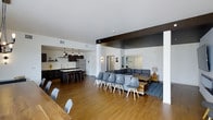 This single-floor space is designed around an open-concept layout that seamlessly connects the kitchen, dining, and lounge areas into one cohesive environment. The kitchen features dark cabinetry, pendant lighting, open shelving, and stainless steel appliances, with a breakfast bar equipped with bar-height seating providing a casual dining option. The kitchen flows naturally into the main living area, where warm hardwood flooring extends throughout, anchoring the space with a sense of continuity and warmth. The lounge area is furnished with a large sectional sofa and a cluster of modern molded chairs arranged around a rustic wooden coffee table, creating a flexible seating arrangement suitable for both small gatherings and larger group settings. A patterned area rug beneath the lounge furniture adds visual texture and helps define the zone within the open floor plan. White walls and a white ceiling keep the space feeling bright and airy, while dark accent beams overhead introduce a bold architectural contrast. Recessed and wall-mounted lighting complement the natural light filtering in through the windows, contributing to a well-lit, inviting atmosphere throughout.