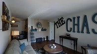 This single-floor home offers a warm and inviting atmosphere, anchored by a thoughtfully decorated living area. The space features rich, dark hardwood flooring that runs throughout, providing a strong visual foundation and a sense of cohesion. The living room is a standout area, showcasing an accent wall with vertical striped paneling that adds texture and depth to the room. Bold decorative lettering adorns the walls, lending the space a personalized, family-centered character. A round wooden coffee table serves as the centerpiece of the seating area, complemented by a patterned area rug and a comfortable sofa dressed with accent pillows. A sleek console table against one wall provides additional display space for framed photos and decorative accents. Wall-mounted shelving displays curated décor items, while a tall decorative floor piece adds vertical interest to the room. The textured white ceiling and neutral wall tones create a bright, airy backdrop that balances the darker flooring. A doorway visible in the background hints at additional rooms, suggesting a practical and connected layout. Overall, the space combines rustic charm with a cozy, lived-in feel that makes it distinctly homey and welcoming.