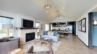 This single-floor home showcases a thoughtfully designed open-concept layout that connects the living, dining, and kitchen areas into one cohesive and welcoming space. The living room is anchored by a clean-lined fireplace with a textured surround, paired with a wall-mounted television, creating a comfortable focal point for relaxation. A light gray sofa, layered rugs, and natural wood accents contribute to the space's warm yet contemporary aesthetic.

Soaring vaulted ceilings with a dramatic peak draw the eye upward, amplifying the sense of spaciousness and airiness throughout the main living area. Two elegant ring-style pendant chandeliers hang from the ceiling, providing both ambient lighting and a decorative touch. Recessed lighting and natural light from multiple windows further brighten the interior.

The kitchen is located at the rear of the open plan and features rich dark cabinetry, a stainless steel refrigerator, pendant lighting over a kitchen island with bar seating, and a tile backsplash. The transition between the living and kitchen areas is seamless, making the layout ideal for entertaining.

Light-toned hardwood flooring runs throughout, tying all areas of the space together cohesively and complementing the neutral color palette. A mini-split climate control unit is also visible, ensuring year-round comfort in this well-appointed home.
