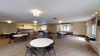 This single-floor event space is designed to comfortably accommodate a variety of gatherings, from meetings and community events to social functions. The room is generously proportioned, offering an open layout furnished with multiple round white tables and black folding chairs arranged throughout, allowing for flexible seating configurations to suit different group sizes.

The interior features warm beige walls accented by dark wood baseboards, contributing to a neutral and welcoming atmosphere. Underfoot, a patterned carpet with an organic, floral-inspired design adds visual interest while defining the main gathering area. Overhead, flush-mount drum-style light fixtures provide consistent ambient lighting across the space.

Natural light filters into the room through several windows fitted with wood-tone Roman shades, offering views of surrounding greenery. Along one side of the room, a counter or service area is visible, suggesting the space may support catered events or refreshment service. The overall ambiance is clean, functional, and adaptable, making this a practical venue for a wide range of organized activities and group gatherings.