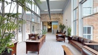 This single-story space showcases a thoughtfully designed interior that seamlessly merges industrial and craftsman aesthetics. The reception or lounge area is anchored by dramatic floor-to-ceiling windows along the perimeter, flooding the space with abundant natural light and offering views of the exterior brick facade. Soaring ceilings with exposed ductwork and structural elements reinforce the industrial character, while warm, variegated brick accent walls introduce texture and visual depth.

Light-toned hardwood flooring grounds the space and complements the rich, dark-stained craftsman-style wooden furniture, including sturdy benches, armchairs, and seating arrangements that invite comfortable gathering. Lush indoor plants — including large tropical varieties and sculptural snake plants — are strategically placed throughout, softening the industrial elements and adding a natural, organic quality to the environment.

A corridor visible through a doorway hints at additional rooms and workspaces beyond, suggesting a larger facility. The overall ambiance balances professionalism with warmth, making the space feel both functional and welcoming for visitors and occupants alike.
