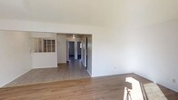 This single-story, 2-bedroom, 1-bathroom residence offers a practical and well-organized layout across a comfortable footprint. The interior features clean white walls throughout, creating a bright and neutral backdrop that lends itself to a variety of décor styles. The main living area is finished with warm-toned hardwood-style flooring, adding a touch of warmth to the open space. Natural light filters into the living area, contributing to an airy atmosphere.

A wide, open passageway connects the living area to the interior hallway, which leads to the bedrooms and bathroom. The hallway is finished with tile-style flooring, providing a durable and easy-to-maintain transition between spaces. A ceiling-mounted light fixture in the hallway ensures consistent illumination throughout. The unit also features a windowed wall detail near the entry area, likely serving as a pass-through or decorative architectural element, adding a subtle point of visual interest.

Overall, the space presents a clean, move-in ready environment with a straightforward single-floor layout, making it a functional and practical living option.