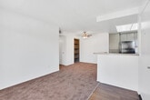 Located on Florence Avenue in Santa Fe Springs, California, this single-floor residence offers a practical and well-organized layout with a clean, neutral aesthetic throughout. The open-concept design connects the living area to the kitchen, creating a cohesive and functional shared space. The living area features plush carpeting and a ceiling fan with a light fixture, while a sliding glass door floods the room with natural light and provides direct access to a private balcony with views of the surrounding landscaped exterior and neighboring tile-roofed structures. The kitchen is equipped with gray cabinetry, granite-style countertops, a full suite of black appliances including a refrigerator, gas range with overhead ventilation, and dishwasher. A skylight brightens the kitchen naturally, complementing the wood-look flooring. A peninsula counter separates the kitchen from the living space, offering a convenient transition zone. The bedroom features neutral carpet, a window with vertical blinds, and a built-in closet with a hanging rod and sliding doors, providing ample storage. The bathroom includes a vanity with granite-style countertops, gray cabinetry, wood-look flooring, and a separate shower area. The entryway transitions seamlessly from hard flooring to carpet, grounding the home's consistent and practical interior design.
