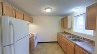 This single-floor unit presents a straightforward and functional layout designed for comfortable day-to-day living. The living area is generously proportioned, featuring neutral-toned carpet underfoot and cream-painted walls that create a bright, clean atmosphere. A textured ceiling adds subtle visual interest throughout the space. Climate control is provided by a wall-mounted air conditioning unit, complemented by baseboard heating along the exterior walls, ensuring year-round comfort. Multiple windows along one wall allow natural light to fill the room, enhancing its open, airy feel.

The kitchen is efficiently arranged in a galley-style configuration, featuring light wood cabinetry that lines both walls and provides substantial storage space. A double-basin sink sits beneath a window, while white appliances—including an electric range and a full-size refrigerator—are integrated seamlessly into the layout. Dark laminate countertops offer a practical workspace, and tile flooring adds durability to the area. Flush-mount ceiling lighting illuminates the kitchen evenly. Overall, this space delivers a clean and practical environment with essential amenities in a well-organized single-level arrangement.