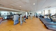 Located at 9200 University City Blvd in Charlotte, NC, this fitness center offers a well-appointed, single-floor exercise facility designed to support a comprehensive workout experience. The space features durable hardwood-style flooring throughout, a drop tile ceiling with recessed lighting, and a clean, bright atmosphere enhanced by natural light streaming through multiple windows along the exterior walls.

The cardio zone is equipped with several treadmills, elliptical trainers, and stationary bikes, positioned to take advantage of the natural light and exterior views. A dedicated strength training area houses a robust selection of Cybex selectorized machines, including cable systems, arm curl stations, chest press units, and seated exercise equipment, providing options for a full-body resistance workout.

Full-length mirrors line portions of the walls, visually expanding the space and supporting proper form during exercise. A standout feature is the vibrant fitness-themed word art mural adorning one wall, adding a motivating and energetic ambiance to the environment. The open floor plan allows for comfortable movement between equipment zones. The facility is well-maintained, thoughtfully organized, and suited for users of varying fitness levels seeking a convenient and complete on-site workout destination.