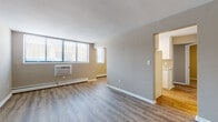 This single-floor unit offers a thoughtfully organized layout across several distinct spaces. The open living area serves as the heart of the unit, featuring durable wood-look vinyl plank flooring, neutral-toned walls, and a textured ceiling. Large windows along one wall allow generous natural light to fill the room, while a wall-mounted air conditioning unit and baseboard heating ensure year-round comfort. The space flows seamlessly toward the kitchen via an open doorway. The galley-style kitchen is efficiently designed with white cabinetry lining both sides, light granite-style countertops, a double basin sink, and white appliances including a range and refrigerator. The kitchen connects directly to the rest of the unit, making it both functional and accessible. The bedroom features the same wood-look plank flooring, a sliding double-door closet providing ample storage, and a neutral color palette. A secondary room is finished with plush carpeting and a window fitted with blinds, offering a quieter, softer environment. The full bathroom is clean and classic, with white square wall tiles in the tub surround, a mosaic tile floor, a toilet, and a vanity. Throughout the unit, consistent warm-toned walls and white trim create a cohesive and welcoming ambiance.