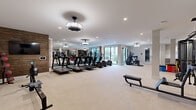 Located in Centennial, CO, this fitness center offers a generously sized, single-floor exercise space designed to accommodate a full spectrum of workout needs. The open floor plan is finished with light-toned flooring throughout, contributing to a clean, contemporary aesthetic that feels both inviting and functional.

The cardio zone features a row of treadmills and elliptical machines positioned near large windows, allowing natural light to flood the space and providing views to the outdoors. A rowing machine adds further variety to the cardio offerings. The strength training area is equally well-equipped, with a multi-station cable machine, squat racks, adjustable benches, and an extensive dumbbell rack spanning a full range of weights, complemented by kettlebell storage. Large wall-mounted mirrors line portions of the room, enhancing the sense of openness and aiding in form monitoring.

Distinctive design elements include a reclaimed wood accent wall that adds warmth and texture against the otherwise neutral, modern palette. Stylish pendant and flush-mount light fixtures supplement recessed ceiling lighting throughout. A wall-mounted flat-screen television and dedicated storage cabinetry add practical amenities. French doors and interior glass partitions connect zones within the space, while emergency exit signage ensures safety compliance. The overall ambiance is polished, spacious, and purpose-built for serious fitness use.