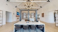 Located at 5401 Commonwealth Centre Pkwy in Midlothian, Virginia, this single-floor space delivers an impressive blend of modern-farmhouse aesthetics and contemporary design. The most striking architectural feature is the soaring vaulted ceiling supported by exposed white timber trusses with bold black metal bracket accents, creating a dramatic sense of height and volume throughout the open interior.

The main gathering area is anchored by a large central lounge featuring warm leather and neutral upholstered seating arranged atop a textured geometric area rug, fostering a welcoming and comfortable atmosphere. A decorative latticed partition with an intricate diamond pattern serves as a refined room divider, elegantly separating the reception desk area from the lounge. The reception island is finished in crisp white and lined with sleek dark bar-height chairs set on a patterned tile inset, adding visual interest underfoot.

Light hardwood flooring flows throughout the space, complementing the bright white wainscoted walls. A floor-to-ceiling open metal shelving unit displays curated décor and greenery, adding warmth and dimension. Natural light pours in through large black-framed windows and glass doors, while recessed and pendant lighting ensures the space is well-illuminated at all times. A cozy nook near the entrance offers additional seating beside expansive windows, ideal for quiet conversation or relaxed waiting.