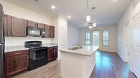 This 1-bedroom, 1-bathroom residence in Austin, TX offers a thoughtfully designed layout that maximizes every square foot. The open-concept kitchen is a standout feature, showcasing rich dark-wood cabinetry, granite-look countertops, a central island with a built-in sink and pendant lighting above, and a full suite of black appliances including a refrigerator, range, microwave, and dishwasher. Recessed lighting and wood-look flooring complete the kitchen's polished look, which flows seamlessly into the living area. The living room is bright and airy, benefiting from large windows and a glass door that invite natural light and provide outdoor views, complemented by a ceiling fan overhead. The bedroom is a comfortable, carpeted retreat with dual windows, a ceiling fan, and a programmable thermostat for year-round comfort. The bathroom features a full tub-shower combination with white tile surround, wood-look flooring, and clean, neutral finishes. A hallway connects the main living spaces efficiently. Throughout the residence, white walls, consistent flooring materials, and modern fixtures create a cohesive, contemporary ambiance well-suited for Austin living.