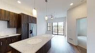 Located in Overland Park, Kansas, this single-floor residence offers a thoughtfully designed layout with contemporary finishes throughout. The open-concept kitchen is a standout feature, showcasing rich dark wood cabinetry that extends to the ceiling, a large quartz-topped island with striking marble-like veining, and a full suite of stainless steel appliances including a refrigerator with ice dispenser, range, microwave, and dishwasher. Elegant pendant lighting and recessed fixtures illuminate the space, while hardwood flooring flows seamlessly through the main living areas. The living area benefits from abundant natural light through large sliding glass doors that open to a private balcony, offering outdoor views of the surrounding neighborhood. A ceiling fan adds both comfort and a finishing touch to the living space. The bedroom features plush carpeting, a ceiling fan, generous window exposure, and access to a walk-in closet, creating a calm and comfortable retreat. The bathroom is appointed with marble-style tile surrounding the soaking tub and shower, a dark wood vanity with a quartz countertop, a large mirror, and wood-look flooring, delivering a refined and cohesive aesthetic throughout the entire residence.