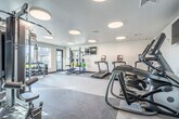 Located at 219 N Fair Oaks Ave in Madison, Wisconsin, this fitness center offers a clean, modern environment designed to support a comprehensive workout experience. The single-floor space is thoughtfully laid out to maximize functionality while maintaining an open, uncluttered feel.

The interior features crisp white walls and a neutral speckled floor, creating a bright, professional atmosphere. A series of large circular flush-mount light fixtures are evenly distributed across the ceiling, providing consistent, well-diffused illumination throughout the space. Multiple flat-screen TVs are mounted on the walls, adding convenience for users during cardio sessions.

The cardio zone is equipped with several treadmills, an elliptical trainer, and a stationary bike, all positioned for easy access. The strength training area includes a Matrix cable machine system, a squat rack with bench, a full dumbbell rack ranging across multiple weights, and additional accessory equipment such as kettlebells, medicine balls, and resistance bands stored on dedicated racks.

Large windows along one wall flood the space with natural light and offer views of an outdoor patio area, enhancing the overall ambiance. A full-length mirror panel reflects the room, contributing to a sense of spaciousness. The fitness center is accessed through a glass-paneled door, adding a sleek, contemporary finish to the overall design.