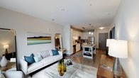 Located at 3310 Women's Club Rd in Raleigh, NC, this single-floor residence offers a thoughtfully designed open-concept layout that connects the living area and kitchen in a cohesive, flowing space. The living room is anchored by a comfortable white sofa, a glass-top coffee table, and a large landscape art piece that adds a pop of color to the neutral walls. Warm hardwood-style flooring runs throughout, lending continuity and a modern aesthetic to the entire space. The kitchen is well-appointed with white shaker-style cabinetry, stainless steel appliances including a refrigerator, stove, and microwave, and a central island with a marble-style countertop that provides additional prep and storage space. Pendant lighting hangs above the island, complementing the track lighting above the kitchen area. A dark gray entry door with sidelight framing provides a defined entryway into the home. The open layout creates a sense of spaciousness and ease of movement between functional areas, making it well-suited for both everyday living and entertaining. Clean lines, neutral tones, and modern fixtures combine to give this residence a polished, contemporary character throughout.