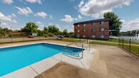 This outdoor pool facility in Merrillville, Indiana, is defined by a generously sized rectangular swimming pool with vivid blue water and a clean concrete deck extending around its perimeter. Stainless steel entry handrails provide safe and convenient access into the water, while depth markers are clearly indicated along the pool's edge. The pool is enclosed by a sturdy wrought iron-style security fence, which runs the full perimeter of the space, providing both safety and defined boundaries for the area.

The surrounding concrete deck offers ample open space for lounging or movement around the pool. A compact utility or storage shed is situated within the enclosure, providing on-site storage for pool-related equipment and supplies. A safety ring is also visible, mounted near the fence line for emergency use.

The facility is set against an open landscape with mature trees lining the background, offering a green, natural backdrop beneath open skies. A multi-story brick residential building is visible adjacent to the pool area, contextualizing this space as a shared outdoor amenity. The overall setting is open and sun-exposed, making it well-suited for recreational use during warmer months.