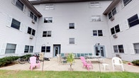This residential building presents a clean, functional design clad in light gray vinyl siding with classic shuttered windows distributed across multiple floors. The exterior is well-maintained, offering a tidy and approachable aesthetic. A standout feature of this space is the ground-level shared patio area, which extends along the rear of the building and serves as a communal outdoor gathering spot. The patio is furnished with a mix of casual seating options, including colorful Adirondack chairs, folding chairs, and tables, creating an inviting atmosphere for residents to relax and socialize. A small potted plant adds a touch of greenery to the hardscaped area. Bicycle storage is conveniently available along the side of the patio, catering to residents who prefer cycling as their mode of transport. Two separate entry doors provide easy access from the patio directly into the building's interior. Window-mounted air conditioning units are visible, ensuring comfort throughout the warmer months. The surrounding lawn area softens the transition between the building and the patio, contributing to the overall livability and charm of this communal outdoor space.