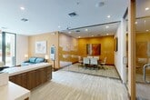 Located at 3400 Davie Rd in Davie, Florida, this well-appointed office space offers a thoughtfully designed, single-floor layout that balances collaborative and individual work environments. The space is anchored by a striking glass-enclosed conference room, providing full transparency while maintaining an acoustic separation from the rest of the office. Inside, a warm-toned accent wall and a large wall-mounted display screen set the stage for productive meetings around a substantial conference table surrounded by ergonomic white chairs. Contemporary artwork adds visual interest throughout. Adjacent to the conference room, a dedicated workstation area features individual computer desks separated by walnut-toned privacy dividers, pendant lighting overhead, and curated artwork on the walls, creating a focused yet creative atmosphere. A comfortable lounge area with a blue sofa, lamp, and framed art offers a welcoming reception or relaxation zone. The flooring consists of patterned carpeting throughout, complemented by recessed lighting in the ceiling that keeps the entire space bright and professional. The open corridor connecting each zone allows for easy navigation, making this office a functional and visually cohesive environment suited for a variety of professional uses.