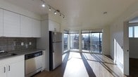This single-floor space showcases a thoughtfully designed open-concept layout that seamlessly integrates the kitchen and living areas. The kitchen is outfitted with contemporary white cabinetry, a stainless steel refrigerator, a dishwasher, and dark countertops complemented by a classic grey subway tile backsplash. Adjustable track lighting runs along the ceiling, providing targeted illumination throughout the cooking and dining space.

The living area benefits from an impressive wall of floor-to-ceiling windows arranged in a corner configuration, flooding the interior with natural light and framing panoramic views of the surrounding cityscape and skyline. The hardwood flooring extends throughout, unifying both spaces and enhancing the sense of openness. The angled window layout creates a distinctive architectural feature that draws the eye outward while maximizing light exposure.

The overall ambiance is clean, modern, and light-filled, with neutral tones and minimalist finishes contributing to a polished, contemporary aesthetic. The elevated position of the unit further enhances the connection to the urban environment beyond.