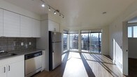 This single-floor space showcases a thoughtfully designed open-concept layout that seamlessly integrates the kitchen and living areas. The kitchen is outfitted with contemporary white cabinetry, a stainless steel refrigerator, a dishwasher, and dark countertops complemented by a classic grey subway tile backsplash. Adjustable track lighting runs along the ceiling, providing targeted illumination throughout the cooking and dining space.

The living area benefits from an impressive wall of floor-to-ceiling windows arranged in a corner configuration, flooding the interior with natural light and framing panoramic views of the surrounding cityscape and skyline. The hardwood flooring extends throughout, unifying both spaces and enhancing the sense of openness. The angled window layout creates a distinctive architectural feature that draws the eye outward while maximizing light exposure.

The overall ambiance is clean, modern, and light-filled, with neutral tones and minimalist finishes contributing to a polished, contemporary aesthetic. The elevated position of the unit further enhances the connection to the urban environment beyond.