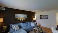 This single-floor space showcases a thoughtfully designed living room that balances bold design choices with a welcoming atmosphere. The most striking feature is a deep chocolate-brown accent wall, which serves as a dramatic backdrop for a large horizontal artwork depicting a illuminated bridge scene, adding a sense of depth and sophistication to the room.

The living area is anchored by a generously sized blue-grey sectional sofa, offering ample seating and a relaxed yet stylish focal point. Decorative throw pillows in complementary tones add texture and visual interest. A round glass-top coffee table sits at the center, reflecting light and contributing to an open, airy feel despite its presence.

The remaining walls are finished in a clean, neutral tone, creating a balanced contrast with the darker accent wall. Warm lighting from a table lamp and wall-mounted light source enhances the cozy ambiance of the room. Hardwood flooring adds warmth underfoot, while framed wall art on the adjacent wall reinforces the curated aesthetic. The overall atmosphere is modern and polished, with carefully selected furnishings and décor contributing to a cohesive, inviting interior.
