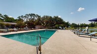 Located at 1718 Northside Dr in Valdosta, Georgia, this outdoor pool area offers a well-appointed recreational amenity designed for relaxation and leisure. The centerpiece is a large, rectangular swimming pool with clear, blue-green water, accessible via stainless steel entry rails. The expansive concrete sun deck surrounds the pool on all sides, providing ample space for sunbathing and socializing. Numerous modern lounge chairs and flat sun benches are arranged throughout the deck, offering plenty of seating for residents and guests. The pool area is fully enclosed by a wrought iron security fence with brick accents, ensuring a safe and private environment. Mature oak and pine trees line the perimeter beyond the fence, creating a lush, natural backdrop that provides partial shade and enhances the serene atmosphere. Additional features visible from the pool deck include a covered playground area and a small utility shed. The open-sky setting allows for abundant natural light throughout the day. The overall ambiance is relaxed and inviting, well-suited for warm Georgia summers, with thoughtful landscaping and greenery contributing to the comfortable, community-oriented character of the space.