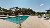 Located at 1718 Northside Dr in Valdosta, Georgia, this outdoor pool area offers a well-appointed recreational amenity designed for relaxation and leisure. The centerpiece is a large, rectangular swimming pool with clear, blue-green water, accessible via stainless steel entry rails. The expansive concrete sun deck surrounds the pool on all sides, providing ample space for sunbathing and socializing. Numerous modern lounge chairs and flat sun benches are arranged throughout the deck, offering plenty of seating for residents and guests. The pool area is fully enclosed by a wrought iron security fence with brick accents, ensuring a safe and private environment. Mature oak and pine trees line the perimeter beyond the fence, creating a lush, natural backdrop that provides partial shade and enhances the serene atmosphere. Additional features visible from the pool deck include a covered playground area and a small utility shed. The open-sky setting allows for abundant natural light throughout the day. The overall ambiance is relaxed and inviting, well-suited for warm Georgia summers, with thoughtful landscaping and greenery contributing to the comfortable, community-oriented character of the space.