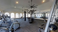 This fitness center offers a comprehensive workout environment designed to accommodate a wide range of exercise needs. The space is anchored by soaring ceilings that create an open, airy atmosphere, enhanced by large arched windows lining the walls and flooding the interior with abundant natural light. Oversized ceiling fans circulate air throughout, maintaining comfort during intense workouts. The flooring features durable, multi-toned carpet tiles suited for high-traffic athletic use. Equipment is thoughtfully arranged to maximize usability, including cardio machines such as treadmills and recumbent bikes, a multi-station cable and weight machine, and a heavy punching bag for boxing and functional training. A full-length wall mirror runs along one side of the room, providing visual feedback during exercises and amplifying the sense of space. Recessed lighting supplements the natural light, ensuring the facility remains well-lit at all times. The overall ambiance is clean and functional, with a layout that allows for easy movement between stations. This single-floor facility is well-suited for both cardiovascular and strength-focused training routines, making it a versatile resource for fitness enthusiasts of varying skill levels.
