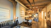 This single-floor loft showcases a striking industrial-chic aesthetic defined by its soaring exposed ceiling with raw ductwork, warm wood beams, and concrete columns that speak to its converted warehouse heritage. The open-concept layout connects the living, dining, and kitchen zones in a fluid, spacious arrangement that maximizes both functionality and visual appeal.

The dining area is anchored by a round wooden table surrounded by sleek, curved white chairs and lit by a geometric pendant light fixture, creating an inviting focal point. A decorative wall display of circular art pieces adds personality and texture to the neutral-toned walls nearby. An industrial-style floor spotlight lamp contributes to the warm, layered lighting throughout.

The kitchen is outfitted with dark cabinetry, stainless steel appliances including a refrigerator, oven, and microwave, and a peninsula with bar seating that doubles as a casual gathering space. Track lighting runs across the ceiling, providing ample illumination across all areas.

Rich hardwood flooring runs throughout, grounding the space and complementing the warm tones of the overhead beams. The living area, partially visible, features a comfortable sofa and curated accessories. Together, these elements create an atmosphere that is both urban and refined, making this loft a distinctive and characterful space.