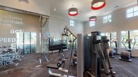This fitness center is a thoughtfully designed, single-level facility offering a comprehensive workout experience. The space is characterized by soaring double-height ceilings and an abundance of natural light streaming through multiple large windows arranged in two tiers along the walls, providing views of lush tropical landscaping and palm trees beyond.

The flooring throughout features a herringbone-patterned carpet tile in deep charcoal tones, lending a modern, sophisticated aesthetic. Bold pendant light fixtures with red accents hang from the ceiling, adding a vibrant design element that complements the otherwise neutral, white-walled interior.

The cardio zone is well-appointed with multiple treadmills, elliptical trainers, a stair climber, and a recumbent bike, all positioned to take advantage of the outdoor views. A dedicated strength training area includes a full rack of dumbbells, a multi-function cable machine, a weight bench, and additional resistance equipment. Stability balls and accessories are neatly stored along one wall.

A distinctive glass partition wall, etched with fitness-themed typography, delineates sections of the space while maintaining an open, airy feel. Wall-mounted mirrors expand the sense of space visually. A small wellness station with shelving rounds out the amenities, making this fitness center a fully functional and inviting environment for a wide range of workout routines.