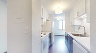 This single-floor unit is thoughtfully organized across several distinct spaces, each finished with practical, clean materials throughout. The galley-style kitchen is a standout feature, designed for efficiency with white upper and lower cabinetry lining both sides of the corridor layout. Dark lower cabinets provide a two-tone contrast, while white countertops, a white electric range and oven, a full-size refrigerator, and drum-style flush-mount ceiling fixtures complete the space. A window at the end of the kitchen draws in natural light, and a baseboard heating unit runs beneath it.

The living areas feature rich, dark hardwood-style flooring that lends warmth to the open layout, while baseboard heaters along the walls provide efficient climate control. Windows allow natural light to filter in, enhancing the bright, neutral palette of white walls throughout.

Bedrooms are carpeted in a neutral tone, with generous windows — one featuring an in-unit wall air conditioning unit — offering outdoor views and ample daylight. Baseboard heating is present in these rooms as well.

The bathroom is finished with patterned tile flooring, a dark vanity with white countertop, a large mirror with overhead lighting, and a full tub-shower combination, providing a clean and functional design.