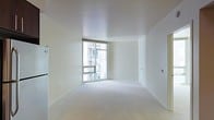 This single-floor space offers a clean, minimalist aesthetic defined by its bright white walls and smooth white ceiling that together create a sense of openness and airiness. The room is finished with neutral-toned wall-to-wall carpeting that adds a soft, understated quality to the overall interior. A standout feature is the large floor-to-ceiling corner window, which allows generous amounts of natural light to flood the space throughout the day. The window provides views of the surrounding urban environment, including neighboring building facades, giving the space a distinctly city-connected character. The walls are largely uninterrupted, offering flexible potential for furniture arrangement and personalization. Subtle architectural details include clean-edged baseboards and smoothly finished wall corners that reinforce the modern, contemporary design language. Electrical outlets and utility panels are positioned at standard intervals along the walls, ensuring practical functionality. The overall ambiance is calm and neutral, well-suited as a blank canvas for a variety of interior styles. The space benefits from excellent brightness and an uncluttered layout that maximizes the sense of scale and livability.