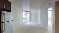 This single-floor space offers a clean, minimalist aesthetic defined by its bright white walls and smooth white ceiling that together create a sense of openness and airiness. The room is finished with neutral-toned wall-to-wall carpeting that adds a soft, understated quality to the overall interior. A standout feature is the large floor-to-ceiling corner window, which allows generous amounts of natural light to flood the space throughout the day. The window provides views of the surrounding urban environment, including neighboring building facades, giving the space a distinctly city-connected character. The walls are largely uninterrupted, offering flexible potential for furniture arrangement and personalization. Subtle architectural details include clean-edged baseboards and smoothly finished wall corners that reinforce the modern, contemporary design language. Electrical outlets and utility panels are positioned at standard intervals along the walls, ensuring practical functionality. The overall ambiance is calm and neutral, well-suited as a blank canvas for a variety of interior styles. The space benefits from excellent brightness and an uncluttered layout that maximizes the sense of scale and livability.