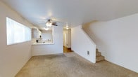 This two-story residence offers a straightforward and functional layout across two levels. The main floor centers around a bright, open living area finished with wall-to-wall carpet, creating a comfortable and cohesive feel throughout the space. A ceiling fan with integrated lighting is mounted overhead, providing both airflow and ambient illumination.

The kitchen is accessible via a pass-through window counter that opens directly into the living area, promoting a connected feel between the two spaces. The kitchen features white cabinetry, a sink positioned beneath the pass-through, and track lighting, keeping the area bright and practical. A hallway visible beyond the living area leads further into the main floor, providing access to additional rooms.

A staircase with clean white walls and carpeted treads rises from the main living area to the second floor, where additional living spaces are located. A wall-mounted heating vent near the base of the stairs ensures climate control throughout the home. The overall aesthetic is clean and neutral, with white walls and light-toned finishes that lend the space a bright, airy quality well-suited for a variety of interior styles. The two-floor configuration provides a natural separation between living and private areas.