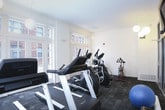 This single-floor fitness center is thoughtfully designed to support a well-rounded exercise routine in a bright, welcoming environment. The space is anchored by an impressive lineup of cardio equipment, including multiple treadmills, elliptical machines, a stair climber, and a stationary spin bike, providing ample options for aerobic training. Large, floor-to-ceiling windows and glass-paneled doors line the walls, flooding the interior with natural light and offering views of the surrounding outdoor environment — creating an open, airy atmosphere despite the compact footprint. The flooring throughout consists of durable, dark-toned carpet tiles well-suited for a high-traffic fitness setting. Overhead, globe pendant lights provide warm, even illumination to complement the natural light. A wall-mounted flat-screen television is installed above a wood-accented shelf near the seating area, where a small cluster of chairs offers a convenient rest space. A glass-partitioned entryway with a full-length mirror connects the main workout area to an adjacent room, adding a sense of openness and depth. The clean white walls and uncluttered layout keep the focus on functionality, making this fitness center a practical and inviting space for daily exercise.