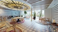 This single-floor communal space combines eclectic design elements with a welcoming, lounge-like atmosphere. The most striking architectural feature is a full-height glass block wall that runs the length of the room, diffusing soft, ambient light throughout the interior while maintaining a sense of privacy. The ceiling is equally distinctive, incorporating inset woven rattan panels framed by white structural beams, lending warmth and texture overhead. A large, ornate circular chandelier serves as a bold focal point, adding an artistic and decorative dimension to the space.

The flooring features a classic two-tone checkered tile pattern in muted gray and beige tones, grounding the room with a timeless yet playful aesthetic. Seating is arranged in intimate clusters around small round tables, each anchored by richly patterned area rugs in warm, jewel-like tones. A built-in wooden shelving unit with a dark countertop lines one wall, functioning as a service or display bar area.

Floor-to-ceiling windows and glass doors along one side open the space to outdoor views and flood the interior with natural light, connecting the inside to a bright exterior setting. Potted tropical plants are positioned throughout, contributing to a fresh, lively ambiance.