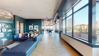 Located atop 131 Mamaroneck Ave in White Plains, NY, this sophisticated amenity floor offers a sweeping, open layout bathed in natural light from floor-to-ceiling windows that frame panoramic views of the White Plains skyline. The space is elegantly designed with a cohesive teal, warm wood, and neutral color palette that flows throughout each zone.

The main lounge area features plush sectional seating, a fireplace with a mounted television, and direct access to an outdoor terrace, creating a seamless indoor-outdoor connection. A separate lounge nook, anchored by a statement crystal chandelier, provides a more intimate setting surrounded by expansive city views on multiple sides.

The gourmet kitchen is a standout feature, showcasing bold teal cabinetry, a large central island with seating, a gas cooktop, and designer lighting—ideal for entertaining. An adjacent private dining area includes a substantial dark wood table beneath pendant lighting, with sheer-draped windows overlooking the cityscape.

A game area with a billiards table adds a recreational dimension, while a dedicated indoor sports simulator room—equipped with a Full Swing system—offers golf and multi-sport simulation. Throughout, warm hardwood and stone tile flooring, exposed ductwork, and curated furnishings contribute to a modern, upscale ambiance.