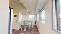 Located at 10045 111 St NW in Edmonton's vibrant Oliver neighbourhood, this rooftop amenity space offers a well-appointed interior combined with generous open-air terraces that deliver sweeping views of the Edmonton skyline. The interior comprises a combined kitchen and living area finished with clean, modern detailing. The kitchen features white flat-panel cabinetry, a taupe quartz countertop, a double-basin stainless steel sink, a glossy tile backsplash, and stainless steel appliances including a full-size refrigerator. Recessed lighting keeps the space bright, while large horizontal windows frame city views and fill the room with natural light. The living area transitions seamlessly from the kitchen, with patterned carpet tiles laid in a herringbone pattern, wall-mounted television, and comfortable seating. A door leads directly from the living area to the outdoor terrace. The rooftop terrace itself is a highlight, offering two distinct zones: an open sun deck furnished with lounge seating, and a covered pergola-style lounge and dining area strung with bistro lighting and equipped with a long communal table and modern white chairs. Glass railing panels ensure unobstructed panoramic views of the Edmonton cityscape throughout.