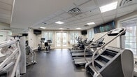 Located at 1 Applewood Drive in Perkasie, Pennsylvania, this fitness center offers a generously sized, single-floor exercise space designed to support a comprehensive workout experience. The room features a drop tile ceiling with recessed and flush-panel lighting that ensures even, bright illumination throughout the space.

The facility is well-appointed with a diverse array of equipment. The cardio zone includes multiple treadmills by Spirit, a stair climber, elliptical trainers, and stationary bikes, providing ample options for aerobic exercise. The strength training area is equipped with a Hoist multi-functional cable machine, a leg press machine, a weight bench, and a cable cross machine, along with a medicine ball rack for functional fitness training.

Durable rubber flooring covers the main exercise areas, transitioning to tile near the entryway. Large windows with blinds along one wall, flanked by French doors, allow natural light to fill the space while offering views of the surrounding greenery. Wall-mounted flat-screen TVs are positioned throughout for entertainment during workouts. Full-length mirrors on one wall enhance the sense of openness and allow for form monitoring. The neutral gray and beige color palette, combined with a pass-through window to an adjacent room, gives the facility a clean, professional, and welcoming atmosphere.